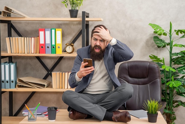 Photo stressed manager reading sms in mobile phone clutching head and sitting crosslegged on desk panic