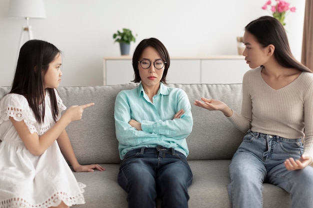 Stressed mature asian woman sitting on couch while adult daughter and granddaughter arguing with her