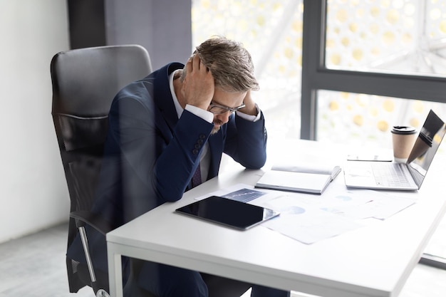 Stressed mature businessman in suit touching head while sitting at workplace