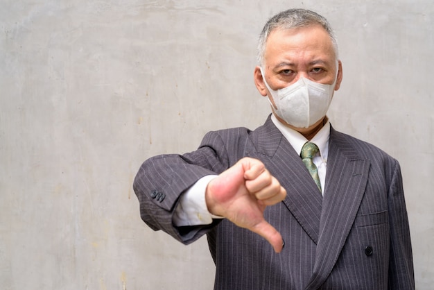 Stressed mature Japanese businessman with mask giving thumbs down outdoors