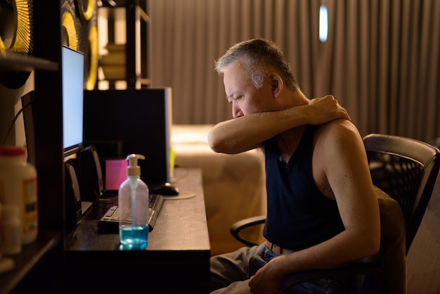 Stressed mature Japanese man covering cough with elbow while working from home