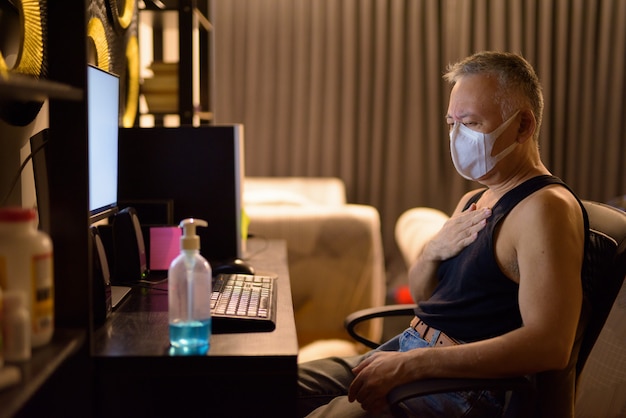 Stressed mature Japanese man with mask getting sick while working overtime at home