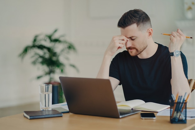 Stressed thoughtful male freelancer or businessman in casual wear feeling tired and having problems at work while sitting at his work desk with laptop at home office, male office worker being nervous