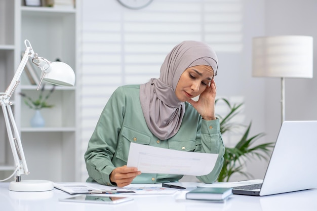 Stressed woman wearing hijab working at home office with laptop and documents experiencing