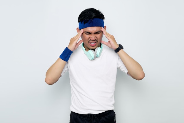 Stressed young Asian sportman in blue headband and sportswear white tshirt with headphonestouching his temples and having headache isolated on white background Workout sport concept