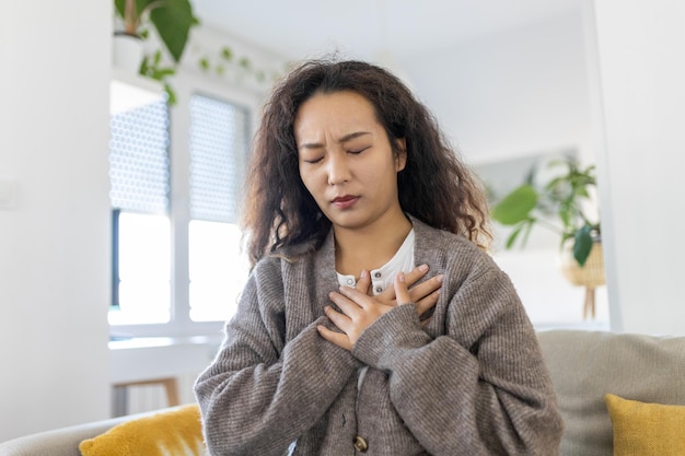 Stressed young Asian woman feeling pain and touching chest suffer from heartache disease at home while having heart attack infarction