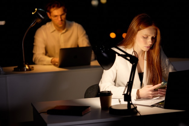 Stressed Young caucasian woman working late night in office late looking sleepy, using smartphone, need some rest, overtime overload deadline working concept. copy space. focus on redhead lady