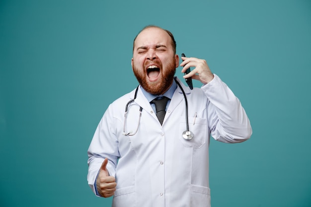 Stressed young male doctor wearing medical coat and stethoscope around his neck talking on phone screaming with closed eyes isolated on blue background