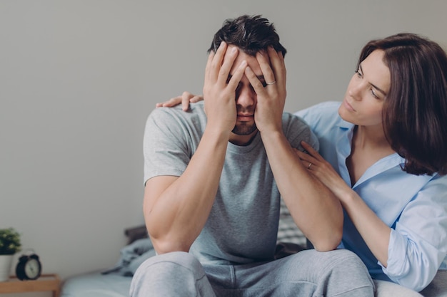 Stressful young man cries from despair loving mother tries to calm him pose together at bed against domestic interior have some problems Upset husband sees no way out in difficult situation