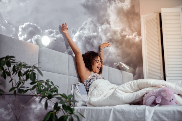 Stretching and yawning. Peaceful dark-haired African American woman wearing pajamas while sitting in bed