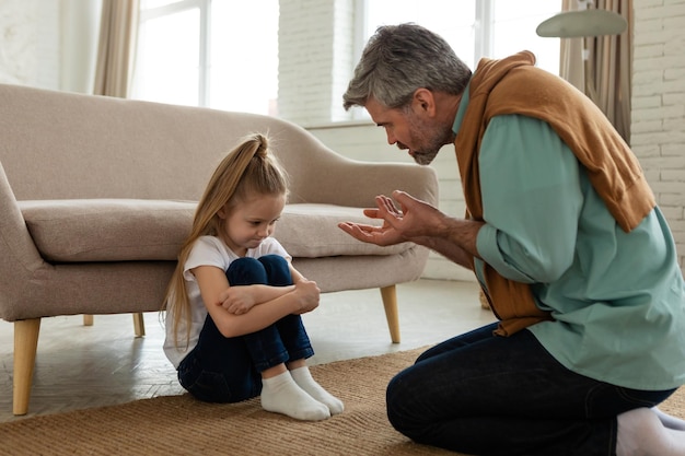 Strict father scolding unhappy daughter sitting on floor at home