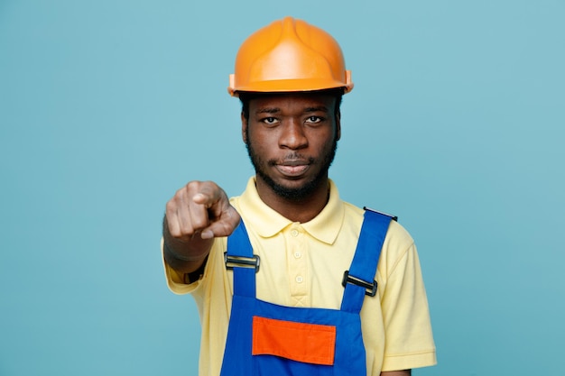 Strict points at camera young african american builder in uniform isolated on blue background