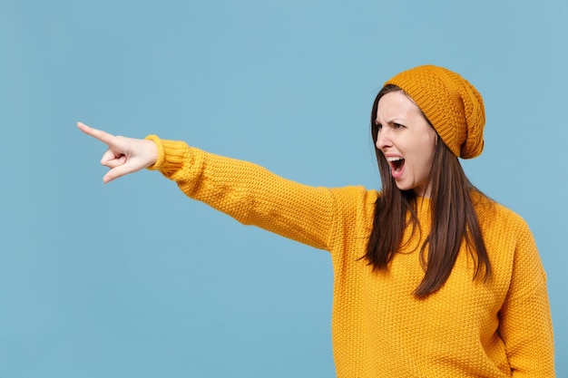 Strict young brunette woman girl in yellow sweater and hat posing isolated on blue wall background studio portrait. People lifestyle concept. Mock up copy space. Pointing index finger aside, swearing.