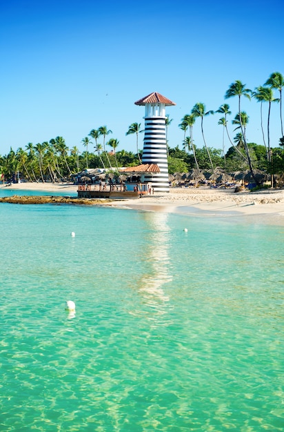 Striped lighthouse on sandy shore with palm trees. Clear water of the Caribbean sea.