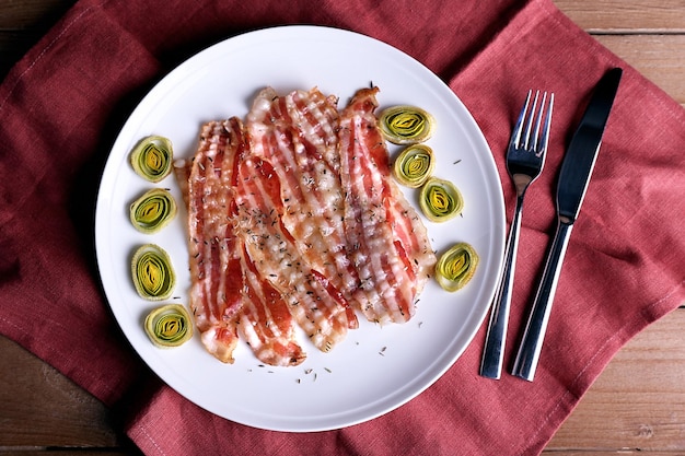 Photo strips of bacon with slices shallot in white plate with napkin on wooden table background