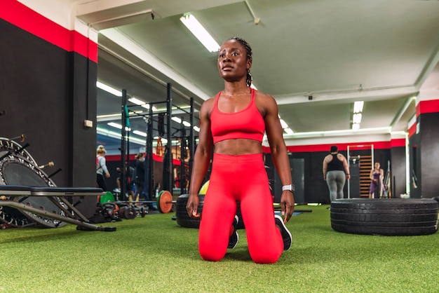 Strong athletic black woman in a gymnasium, ready and concentrating on her knees to do sports, dressed in red sportswear