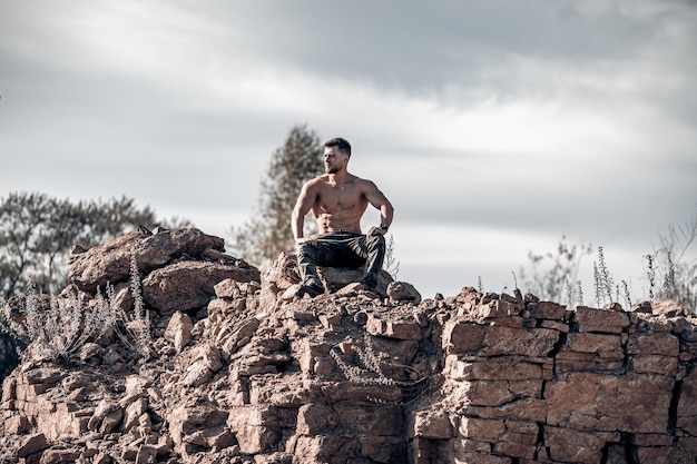 Strong bearded man in military pants Bodybuilder sitting on a rock and looking away Naked torso Full length view