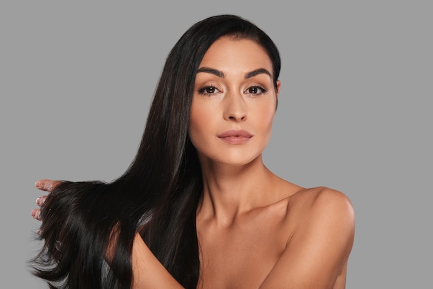 Strong and beautiful hair. Attractive young woman looking at camera with a smile while standing against grey background