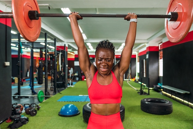 Strong black athlete in a gymnasium doing barbell and weightlifting exercises to strengthen shoulders and biceps showing a lot of effort and in red sportswear