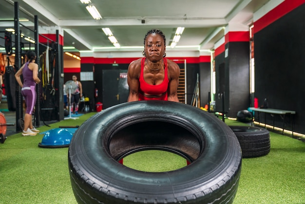 Strong black female athlete in a gym doing wheel lifting exercises to strengthen quadriceps and biceps in red sportswear