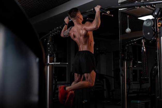 Strong bodybuilder man in good physical shape and a naked muscular body pulls up in the sports gym against a dark background