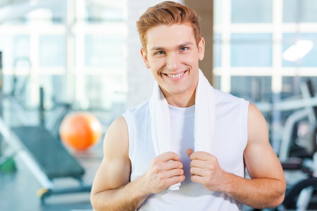 Strong and confident. Confident young muscular man carrying towel on shoulders and smiling while standing in gym