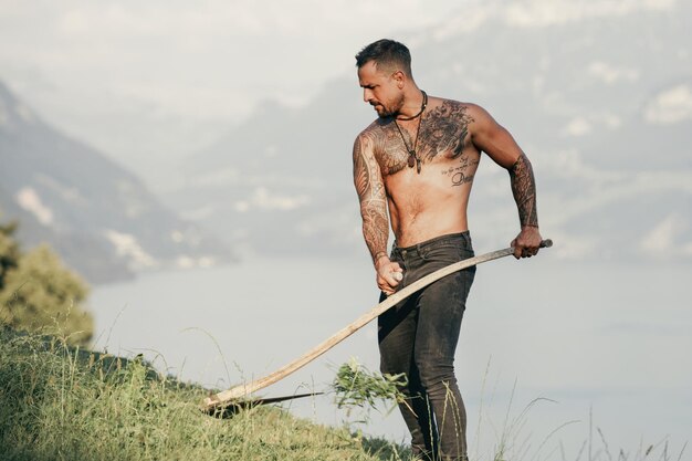 Strong farmer using scythe sexy farmer with a scythe on green grass field muscular farmer cut grass