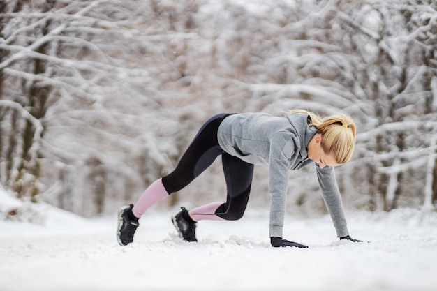 Strong fit sportswoman doing warmup exercises while standing in snow in nature at winter. Healthy lifestyle, winter fitness