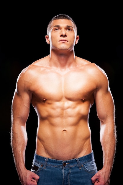 Strong man. Handsome young shirtless man looking up while standing against black background