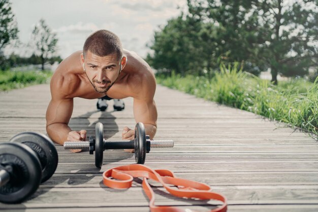 Strong motivated handsome man stands in plank pose makes abdominal exercises poses near sport equipment listens music in wireless earphones has workout outdoor looks with serious expression