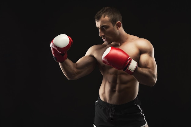 Strong muscular man in red boxing gloves at black studio background, low key, copy space