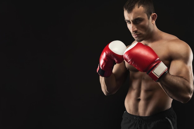 Strong muscular man in red boxing gloves at black studio background, low key, copy space