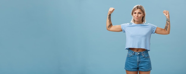 Strong and powerful goodlooking sportswomen with tattoos in tshirt and shorts raising arms showing big muscles and biceps smiling proudly while bragging with physical strength over blue wall