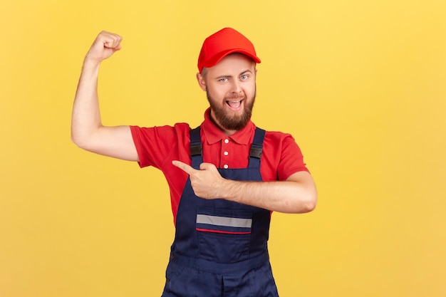 Photo strong worker man wearing blue uniform and red cap standing with raised arms and pointing at biceps