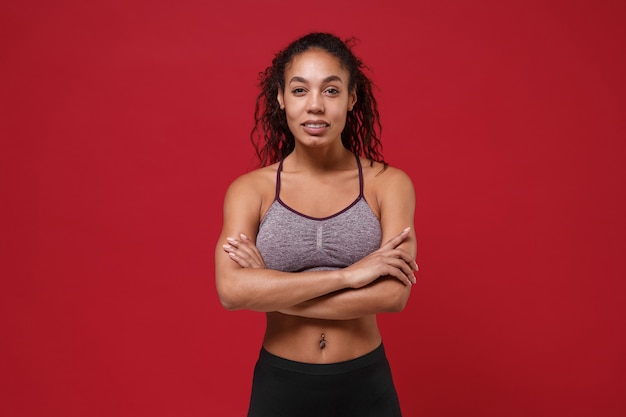 Strong young african american sports fitness woman in sportswear posing working out isolated on red wall  portrait. Sport exercises healthy lifestyle concept. Holding hands crossed.