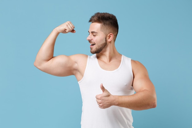 Strong young bearded guy 20s in white singlet posing isolated on pastel blue background studio portrait. Sport fitness healthy lifestyle concept. Mock up copy space. Showing biceps muscles, thumb up.