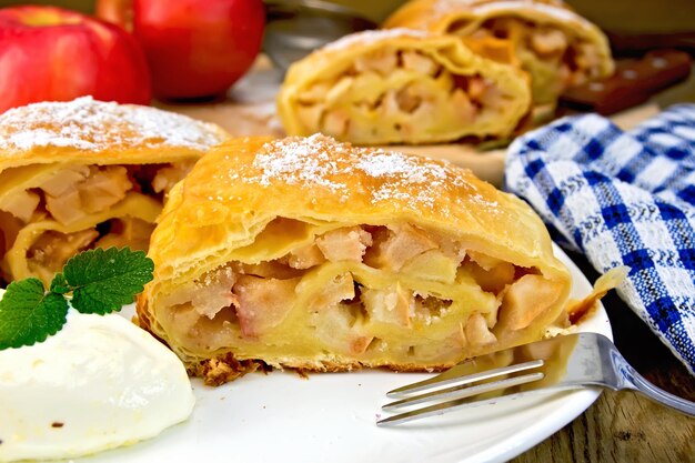 Strudel with apples and ice cream in a white plate with a fork apples knife napkin on the background of wooden boards