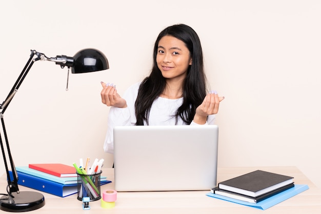 Student asian girl in a workplace with a laptop on beige wall making money gesture