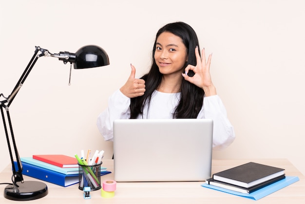 Student asian girl in a workplace with a laptop on beige wall showing ok sign and thumb up gesture