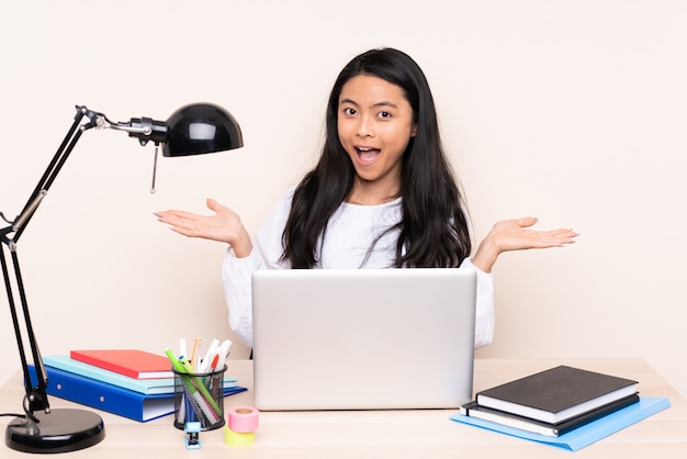 Student asian girl in a workplace with a laptop isolated on beige background with shocked facial expression