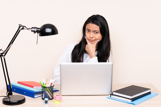 Student asian girl in a workplace with a laptop isolated on beige thinking