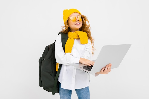 Student girl in casual clothes with a backpack and with a laptop in her hands walking on a white wall