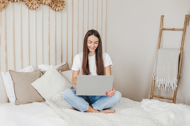 Student girl sits on the bed with a laptop looks at the monitor and smiles