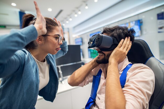 student girl teasing to her bearded boyfriend while testing VR in a tech store.