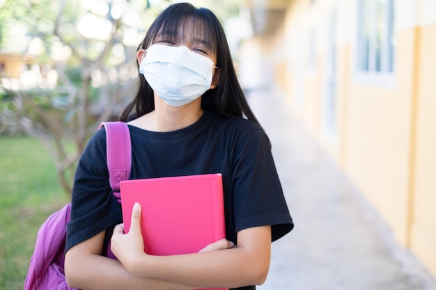 STudent girl wear mask holding book and backpack at school.