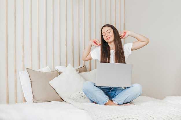 Student girl woke up stretches and sits with a laptop on the bed going to work online