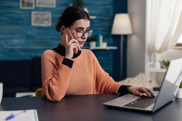 Student at home talking on the phone while using laptop. Programmer with glasses working from home using smartphone and browsing computer. Focused freelancer using technology for remote work.