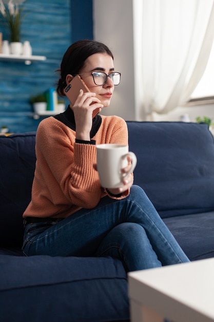 Student talking with her friends about social media using modern smartphone, relaxing alone on couch during coronavirus quarantine. Teenager gossiping while drinking coffee