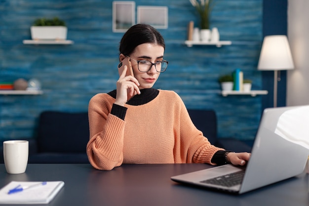 Student working at homework for college seminar using laptop computer browsing information on internet. Young woman talking on smartphone while sitting at table desk in living room