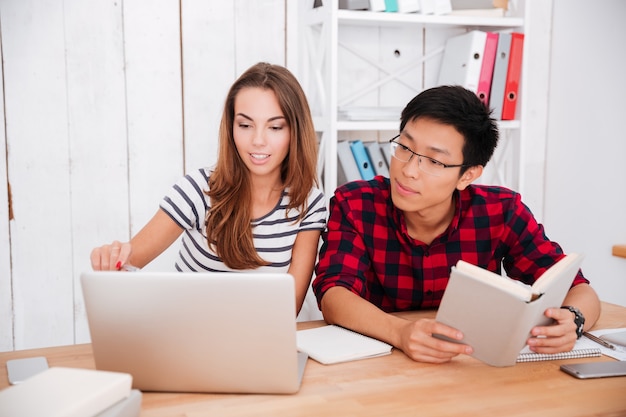 The students are both sitting beside each other and smiling as they use the laptop as woman points to boy on screen